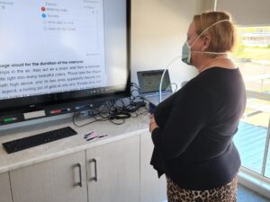 Aged care worker undergoing a quantitative mask fit test at Palm Lake Care Logan Reserve, reading text aloud from a large screen while wearing an N95 respirator connected to sampling tubing.